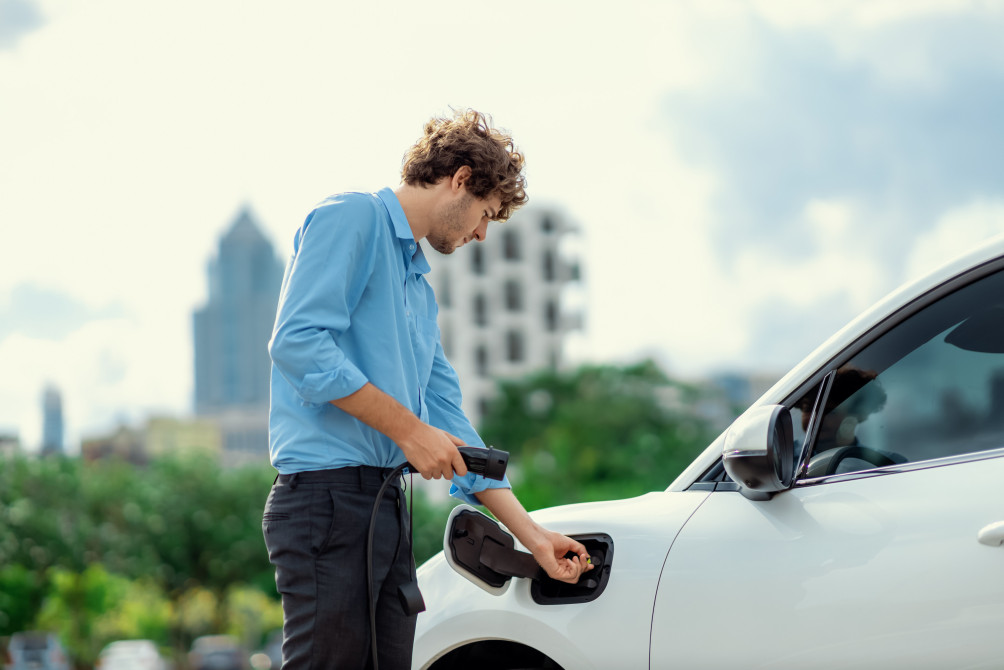 Progressive businessman insert charger plug from charging station to his electric vehicle with apartment condo building in background. Eco friendly rechargeable car powered by sustainable energy.
